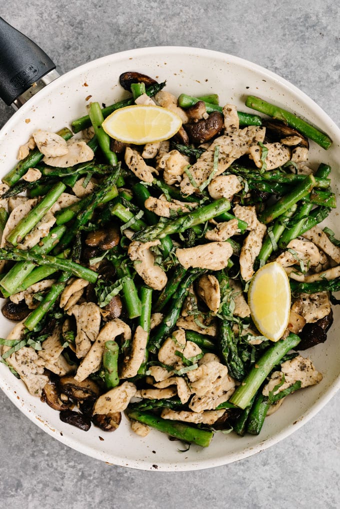 Lemon garlic chicken and asparagus recipe in a skillet with lemon wedges and fresh basil.
