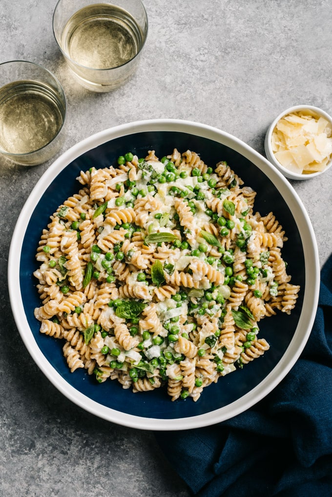 A large serving bowl of ricotta pasta with peas and sautéed leeks with two glasses of white wine and a small side of shaved parmesan cheese.