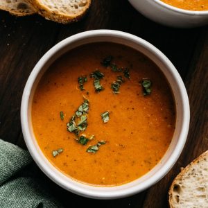 A bowl of roasted tomato soup garnished with fresh basil with slices of sourdough bread on the side.
