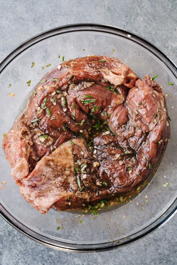 Raw flank steak in a large mixing bowl marinating with rosemary, olive oil, balsamic vinegar, garlic, salt, and pepper.