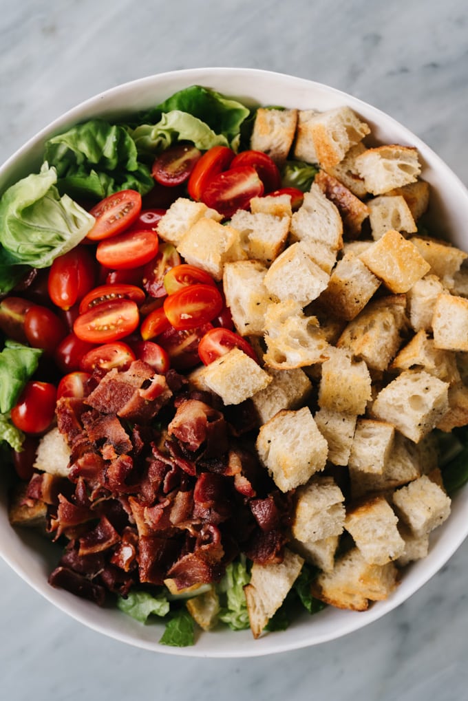 From above, a white salad bowl filled with the ingredients for a BLT salad - chopped lettuce, grape tomatoes, bacon, and sourdough croutons.
