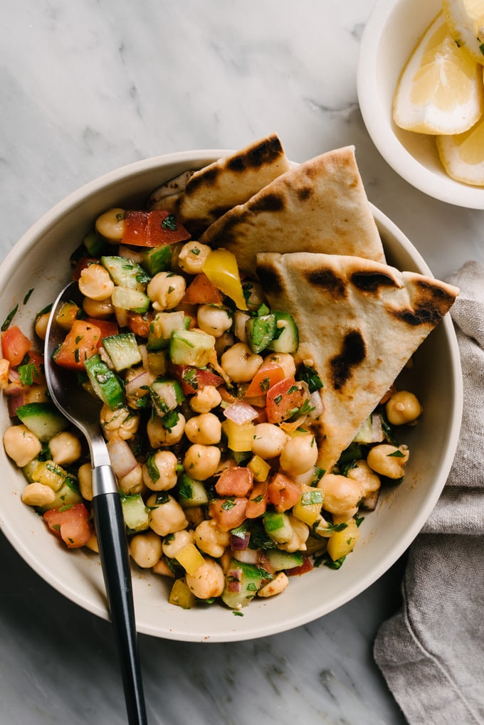 A bowl of mediterranean chickpea salad with slices of pita, a small bowl of lemon slices, and a linen napkin.