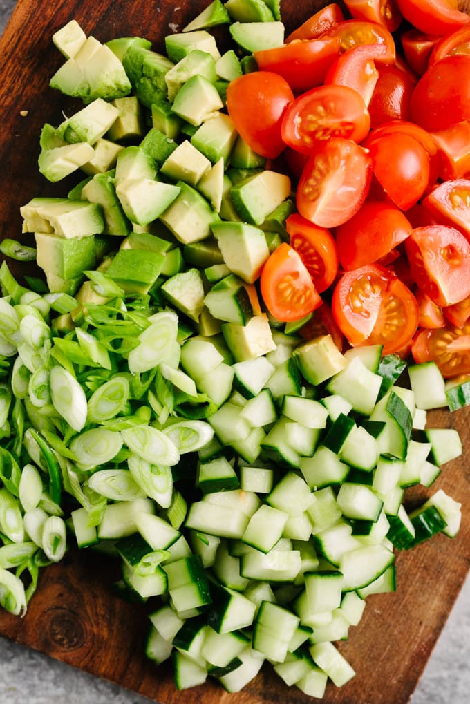 Overhead view of diced avocado, cucumber, tomatoes, and sliced green onions on a cutting board.