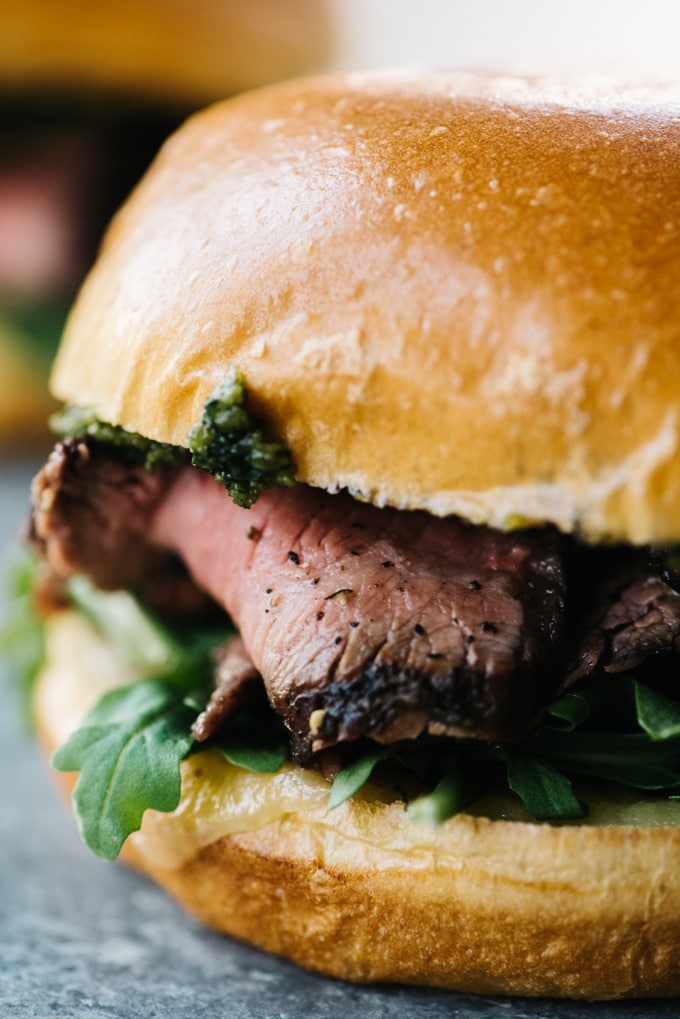 A close-up view of a grilled steak sandwich with sharp cheddar, arugula, thinly sliced steak, and pesto on a brioche bun.