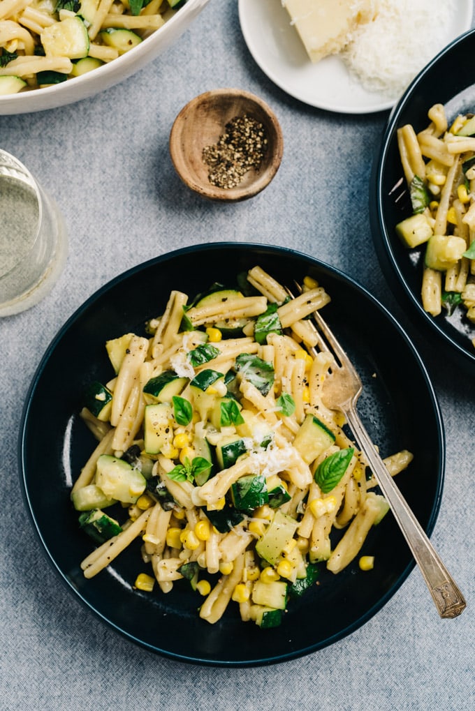 Two bowls of summer pasta with zucchini and sweet corn on a table with a glass of wine and side of parmesan cheese.