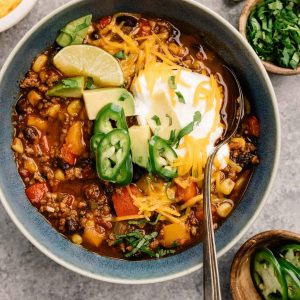A bowl of weeknight ground beef taco soup surrounded by garnishes on a cement background.