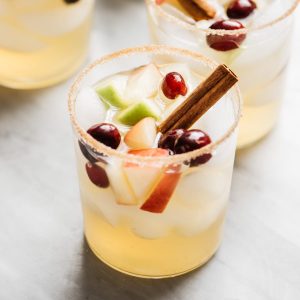 Apple cider sangria cocktails with a cinnamon sugar rim on a marble table with a pitcher of sangria and a smaller jar of cinnamon sticks.
