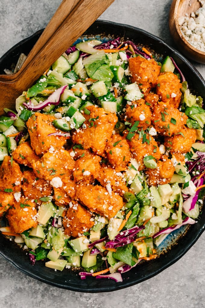 Buffalo chicken salad in a large salad bowl with boneless buffalo wings, with a bowl of blue cheese crumbles to the side.