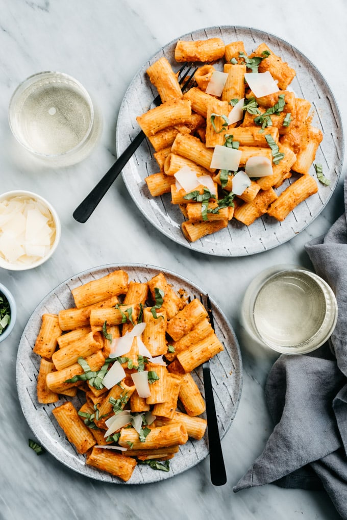 Two plates of rigatoni tossed in creamy tomato sauce on a marble table with white wine and a small bowl of parmesan cheese.