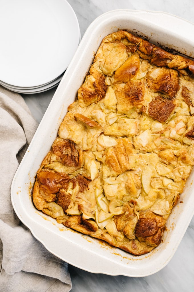 Apple bread pudding in a casserole dish on marble table alongside a stack of white plates and a linen napkin.