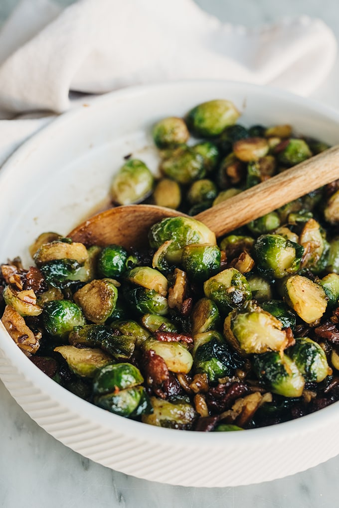 Side view, bacon Brussels sprouts tossed with maple syrup and pecans in a white bowl with a wood spoon.