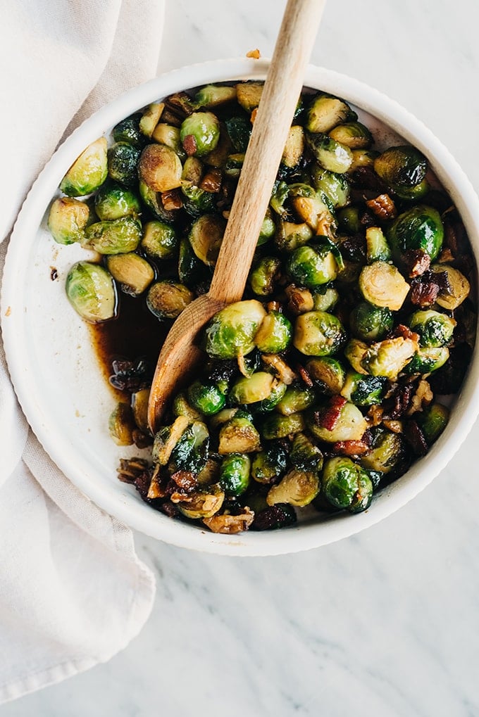 A bowl of maple bacon Brussels sprouts on a marble table with a wood spoon and cream linen napkin to the side.