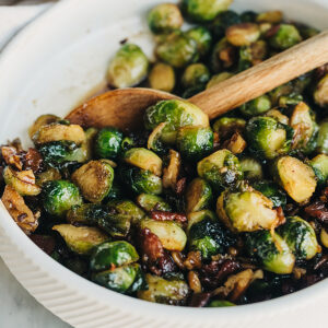 Side view, bacon Brussels sprouts tossed with maple syrup and pecans in a white bowl with a wood spoon.