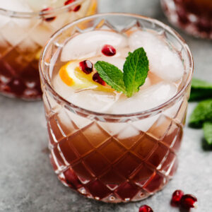Side view, a prosecco punch cocktail garnished with pomegranate seeds, orange slices, and a fresh mint sprig on a concrete table.