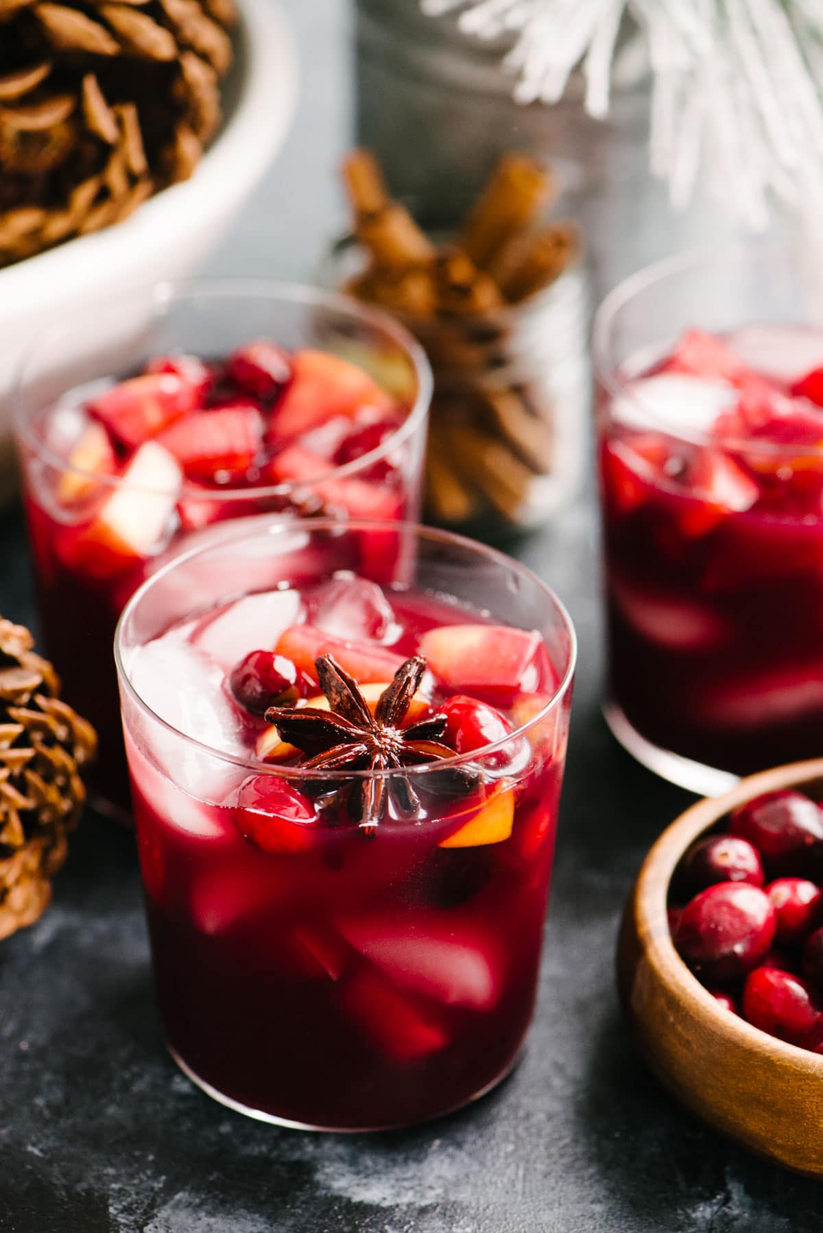 Three glasses of christmas sangria on a black tabletop surrounded by pine cones and a small decorative tree.