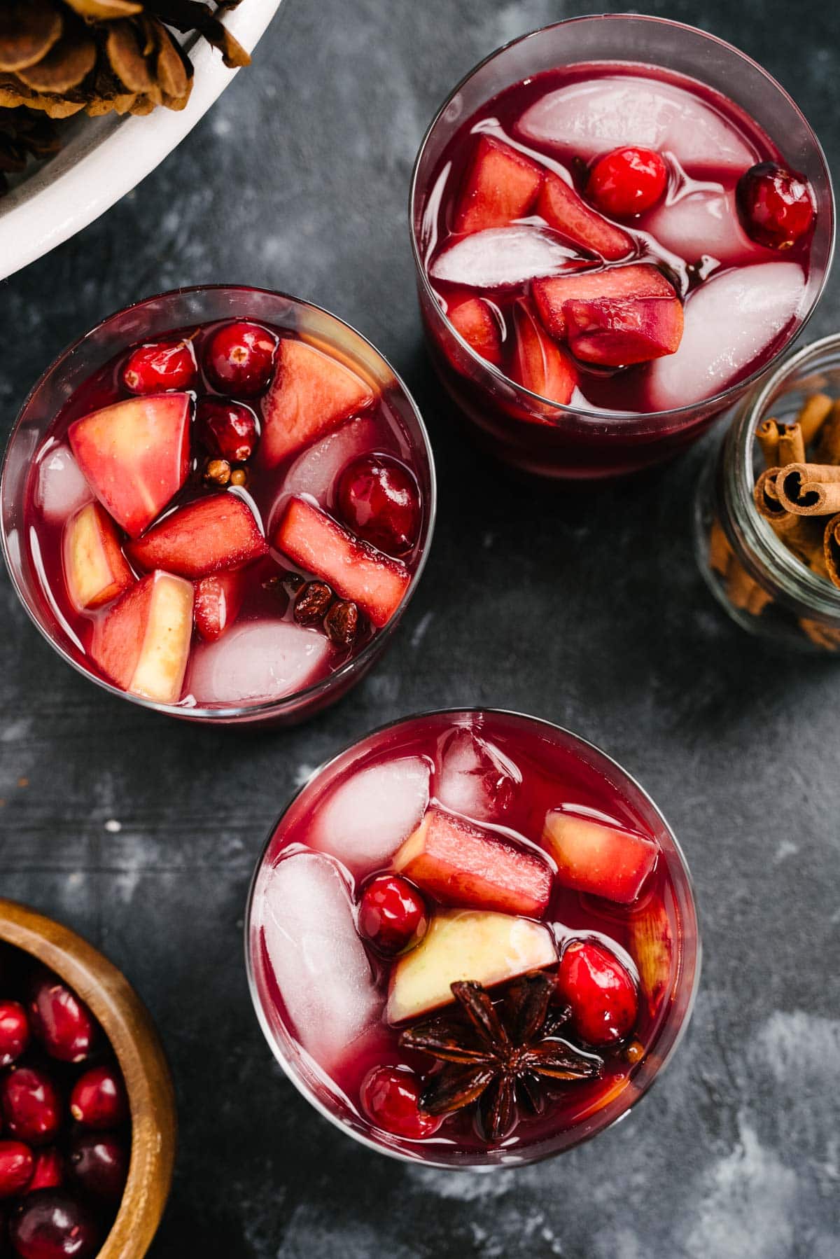 From overhead, three glasses of red wine holiday sangria on a black chalk background with a small wood bowl of cranberries and a small glass jar of cinnamon sticks to the sides.