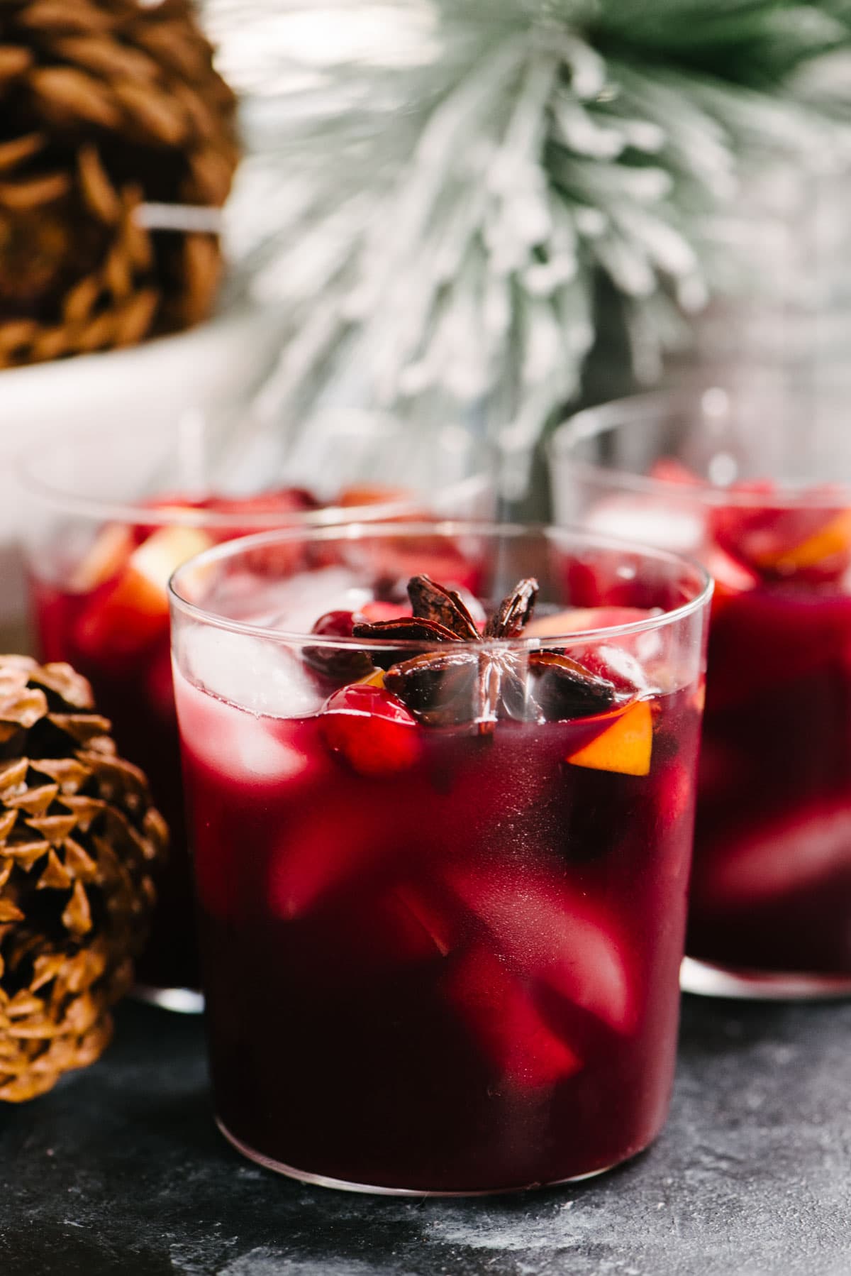 Side view, several glasses of red wine Christmas sangria on black charcoal table with a bowl of pinecones and faux Christmas tree in the background.
