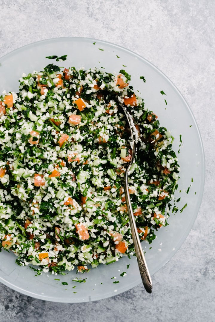 Overhead, grain free cauliflower tabbouleh in a glass serving bowl with a silver spoon.