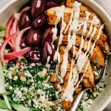 Chicken shawarma bowl with tabbouleh, olives, pickled red onions, and tahini dressing on a cement background with a tan linen napkin.