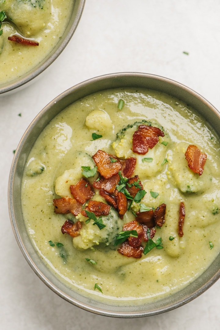 A bowl of broccoli potato soup garnished with crispy bacon on a cement background.