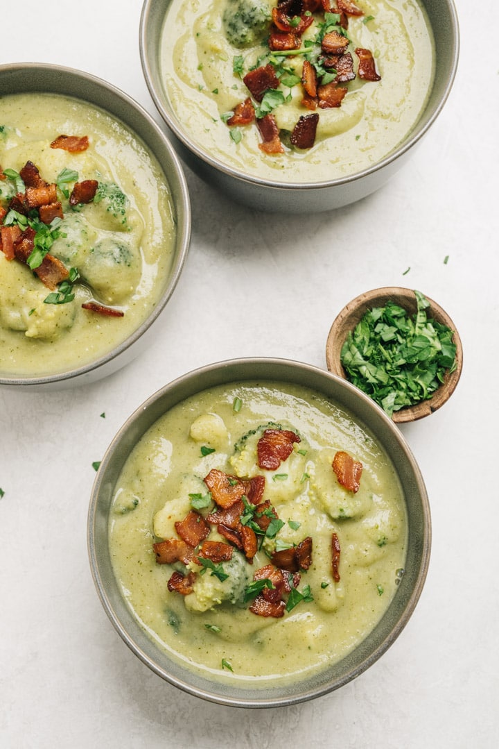 Three bowls of creamy broccoli soup garnished with bacon and chopped parsley.