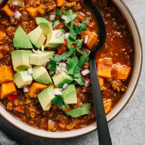A black spoon tucked into a bowl of healthy chili with ground beef and sweet potatoes, topped with avocado, cilantro and diced red onions.