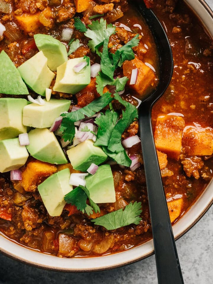 A black spoon tucked into a bowl of healthy chili with ground beef and sweet potatoes, topped with avocado, cilantro and diced red onions.