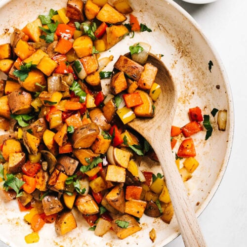 A wood spoon into a skillet of quick and easy sweet potato hash, with a small bowl of chopped parsley to the side.