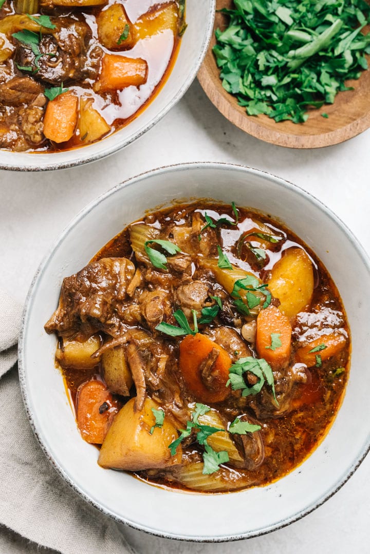 Two bowls of beef stew on a cement background with a pinch bowl of fresh chopped parsley.