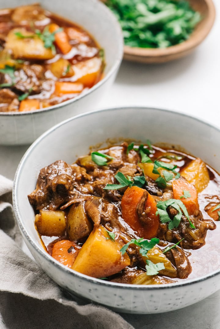 Side view, several bowls of dutch oven beef stew in white bowls on a cement background with a small pinch bowl of chopped parsley and tan linen napkin.