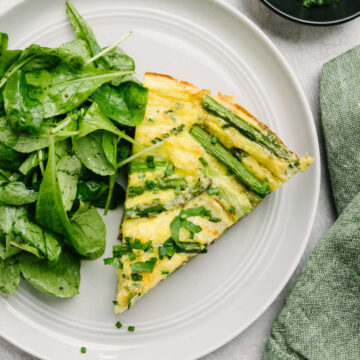 A slice of asparagus frittata on a grey plate with a tossed green salad; a bowl of chopped parsley and a green linen napkin surround the plate.