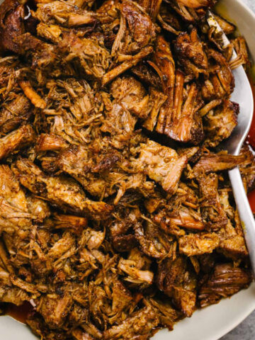 A silver serving fork tucked into a bowl of Instant Pot pulled pork, drizzled with pan sauce.