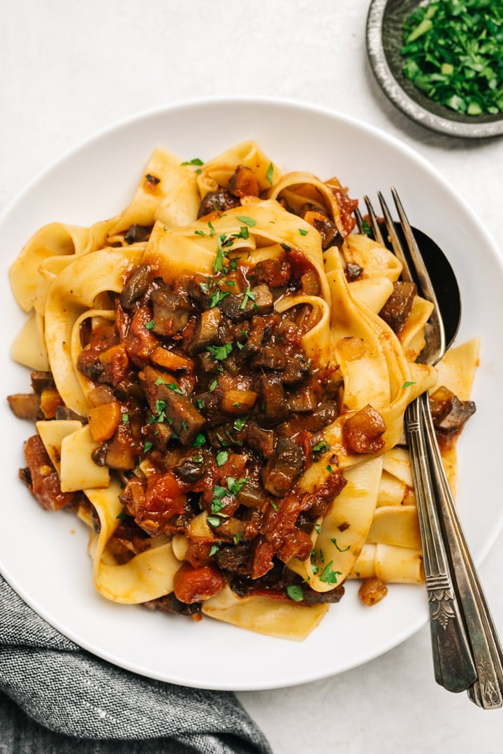 Mushroom bolognese over pappardelle noodles in a white pasta bowl with a vintage fork and spoon, small bowl of chopped parsley, and dark grey linen napkin.