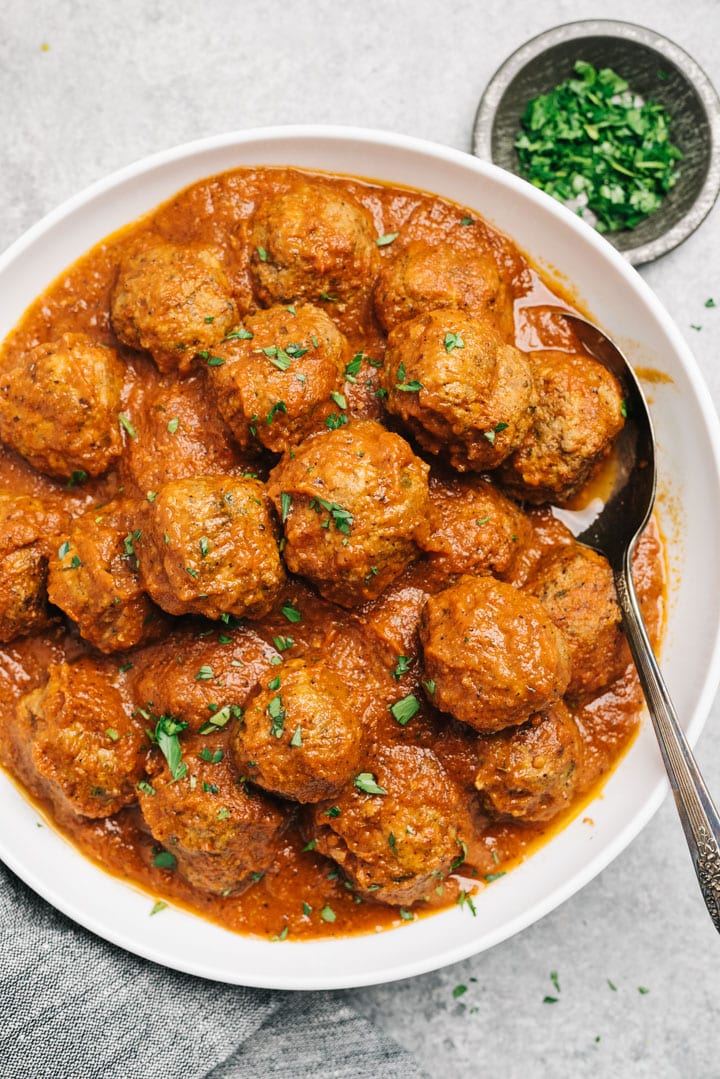 Healthy italian style meatballs tossed with marinara sauce in a large white serving bowl with a small bowl of chopped basil to the side.