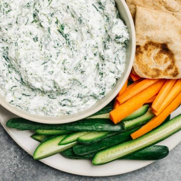 A bowl of healthy greek yogurt spinach dip surrounded by cut vegetables and pita bread.