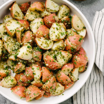 Italian potato salad tossed with an herb vinaigrette in a large white serving bowl with a striped linen napkin tucked to the side.