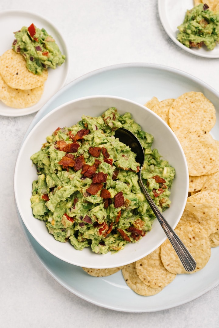 A bowl of bacon guacamole surrounded with chips and two serving plates with chips and guacamole.