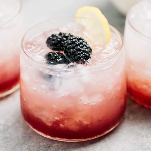 Three gin and blackberry cocktails on a cement surface with small bowls of fresh blackberries and lemon wedges.