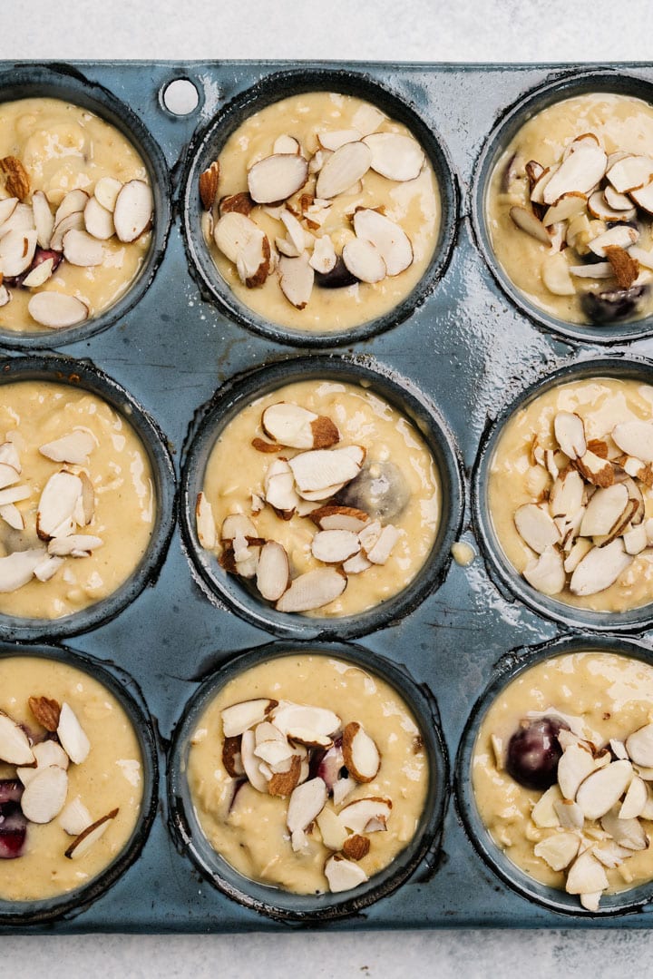 Cherry muffin batter in a muffin tin, topped with sliced almonds.