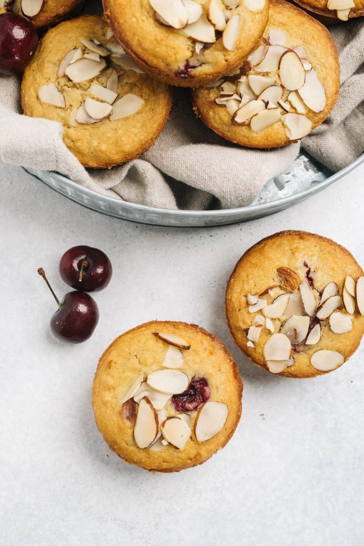 Two cherry almond muffins on a table next to a large bowl of muffins.