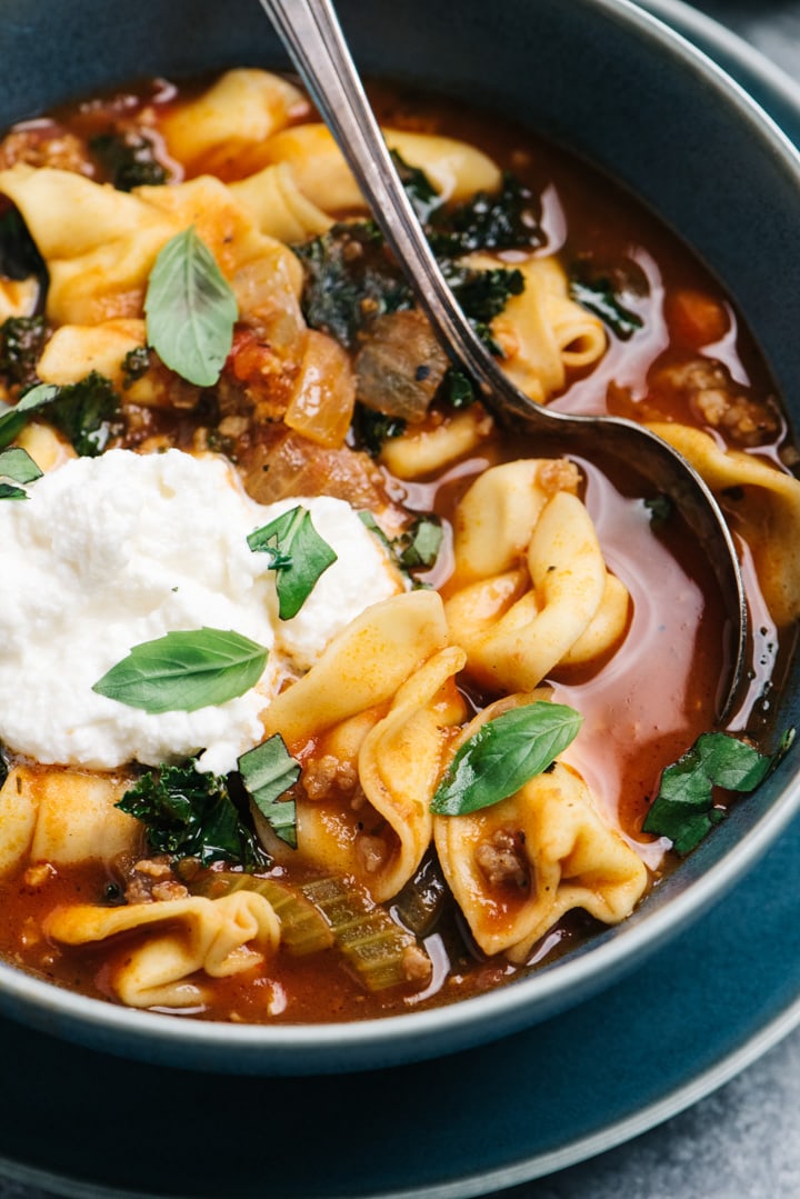 Side view, sausage tortellini soup with kale in a blue bowl with a silver soup spoon.