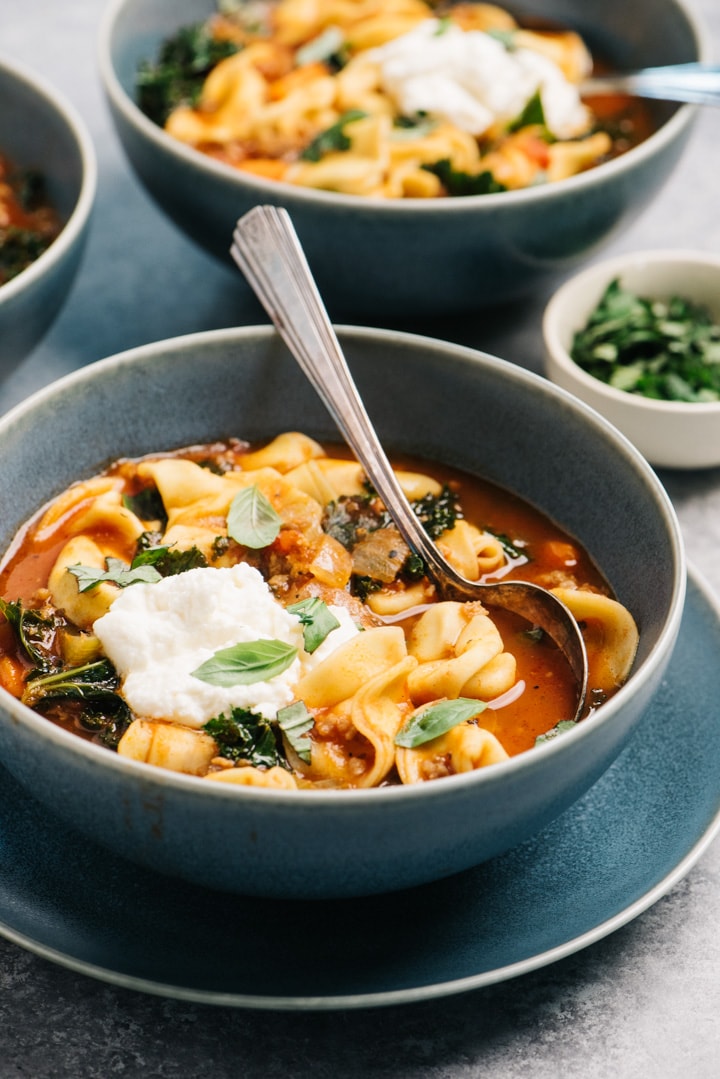 Side view, a bowl of italian sausage and tortellini soup with kale, fresh ricotta, and basil. 