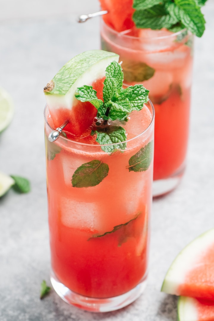 Two watermelon mojito cocktails on a cement background with watermelon wedges, limes, and fresh mint leaves.