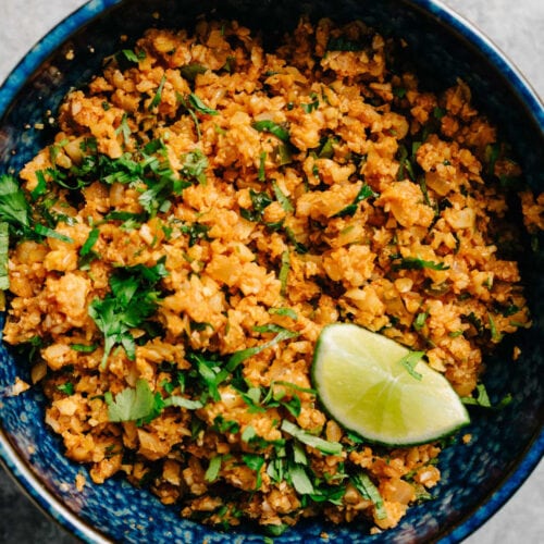 Mexican cauliflower rice in a blue serving bowl on a concrete background, garnished with chopped cilantro and a lime wedge.