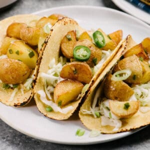 Side view, three potato tacos with serrano chilies on a white plate, garnished with coleslaw and green onions.
