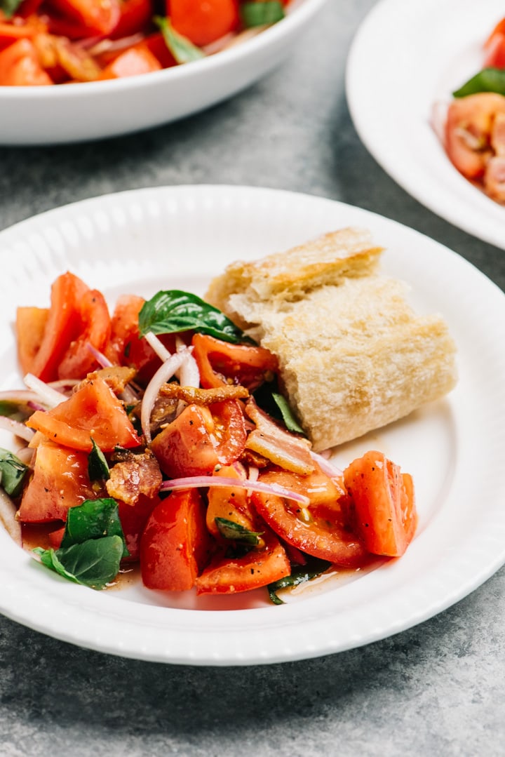Tomato salad with bacon dressing on a white plate with a slice of crusty bread for dipping.