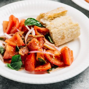 Tomato salad with bacon dressing on a white plate with a slice of crusty bread for dipping.