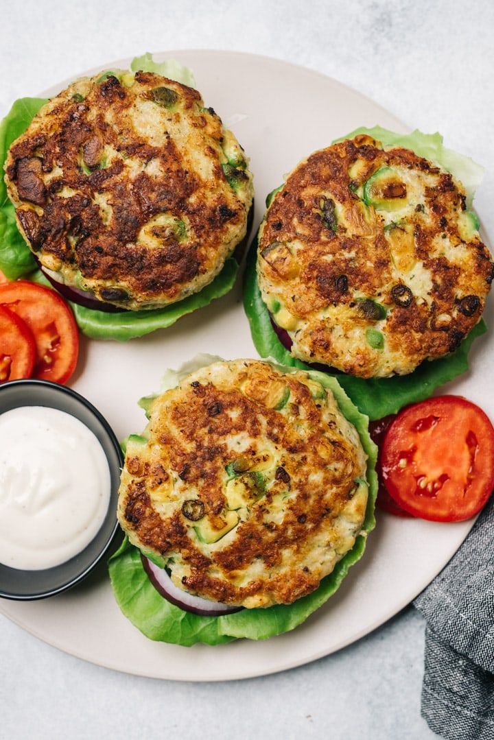 Three ranch chicken burgers with avocado arranged on a plate with lettuce, tomato, onion, and a small dish of ranch dressing.