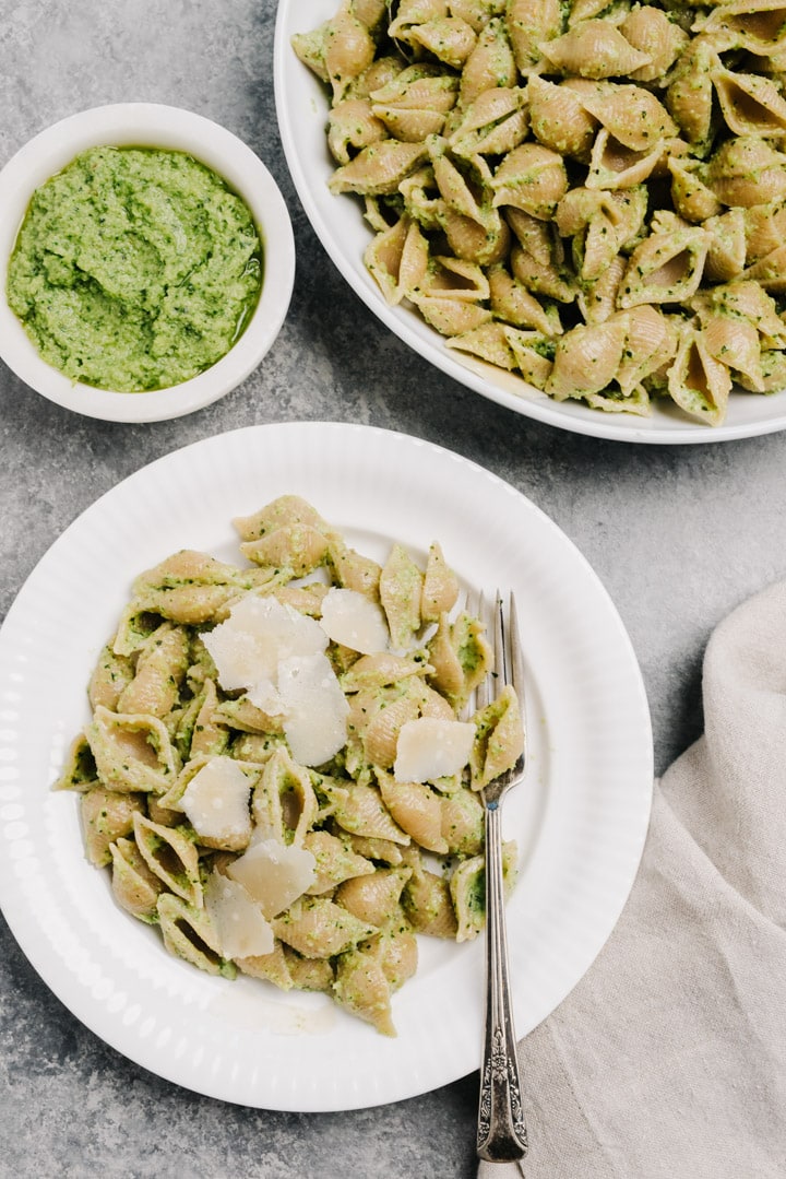 A serving of broccoli pesto pasta on a white plate with a large serving bowl of pasta and a small bowl of pesto on a cement table.