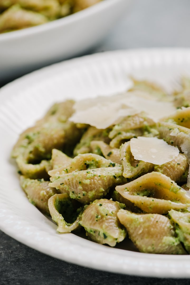Side view, whole wheat shells tossed with broccoli pesto on a white plate, garnished with shaved parmesan cheese.